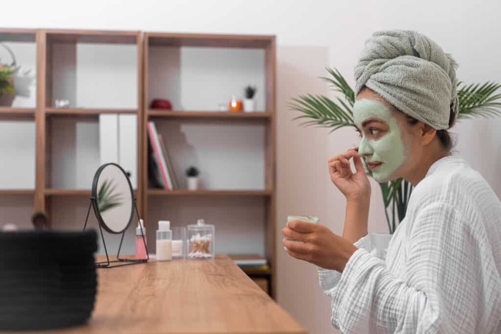 Concentrated woman applying konjac alcohol mixed clay mask on face looking in table mirror pretty lady in bathrobe cleansing face with organic cosmetics at home side view