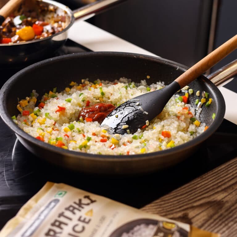 A black frying pan on a stovetop contains shirataki rice being stir-fried with colorful vegetables.