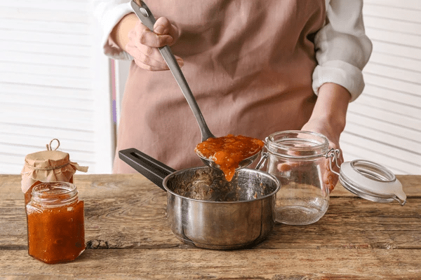 A person ladling a bright orange preserve, a fruit compote for a konjac jelly recipe, into a jar.