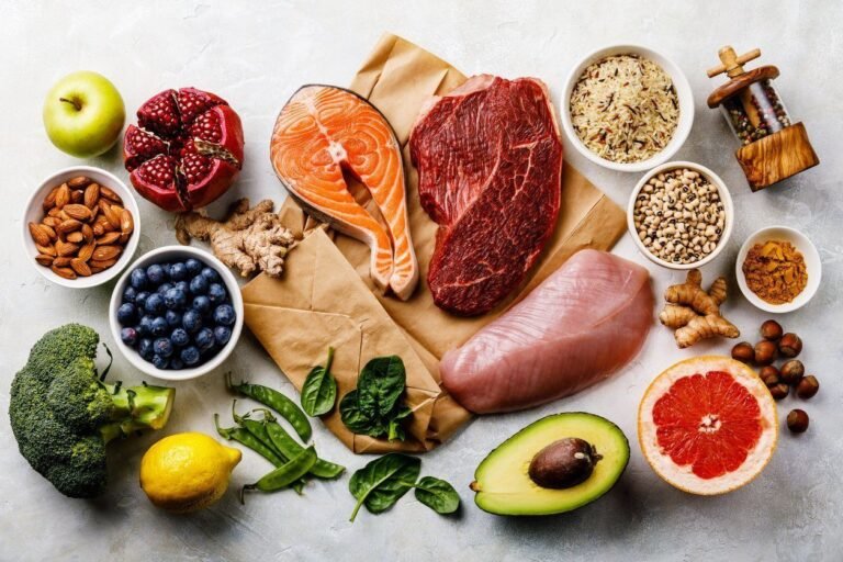 Healthy foods for a balanced diet - salmon, steak, chicken, fruits, vegetables, nuts, and grains on a gray background.