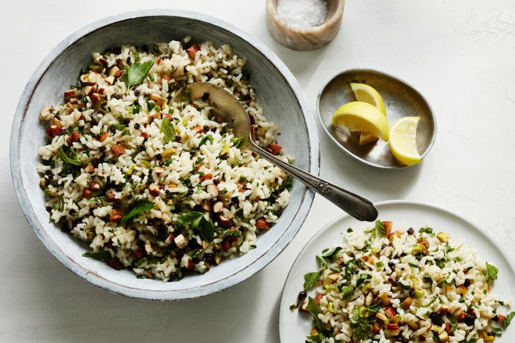 A bowl and plate filled with a refreshing rice salad, garnished with fresh herbs, sliced almonds, and pistachios with lemon wedges beside.