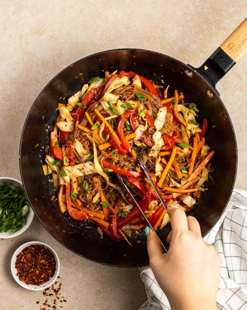 A black wok filled with a colorful stir-fry of konjac FODMAP noodles, carrots, bell peppers, cabbage, and onions, and a hand using tongs to toss the ingredients.